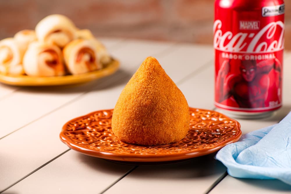 Coxinha em prato laranja, com enroladinhos e refrigerante.