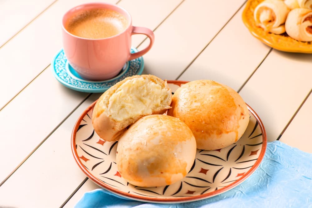 Pães de queijo com requeijão, com café em xícara rosa.