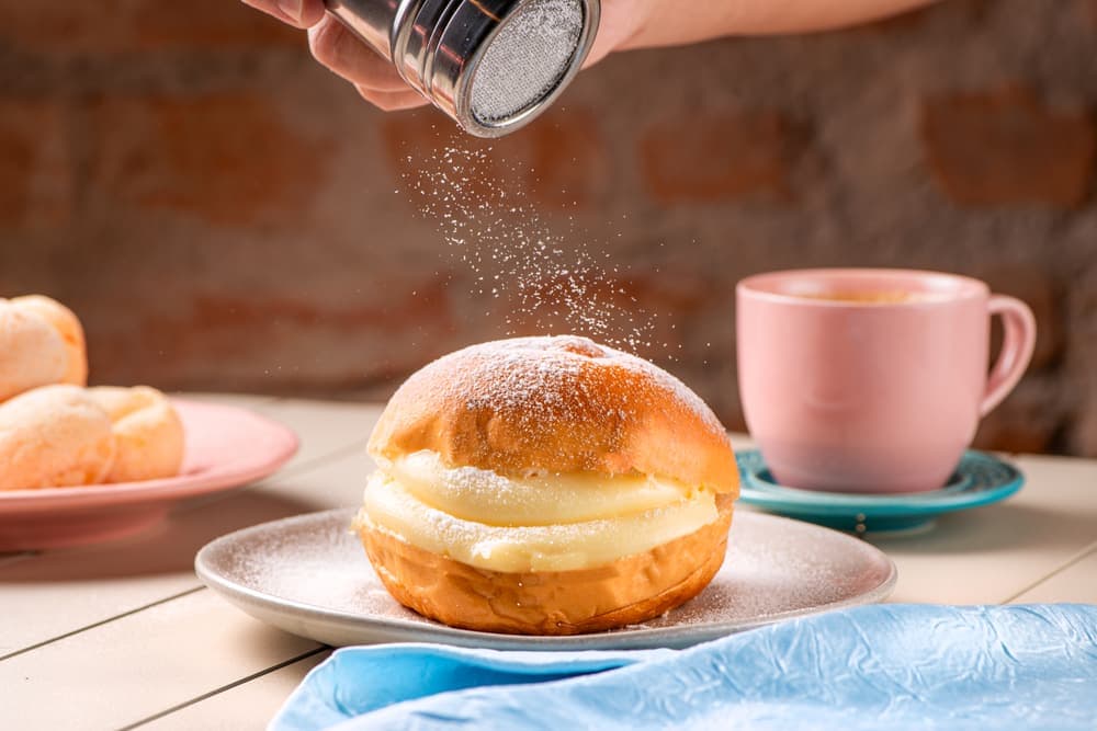 Sonho de creme sendo polvilhado de açúcar, com café.