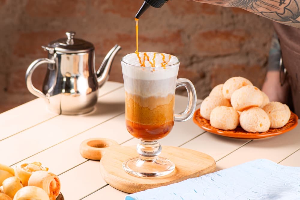 Café com leite em taça de vidro, com bule e pães de queijo.
