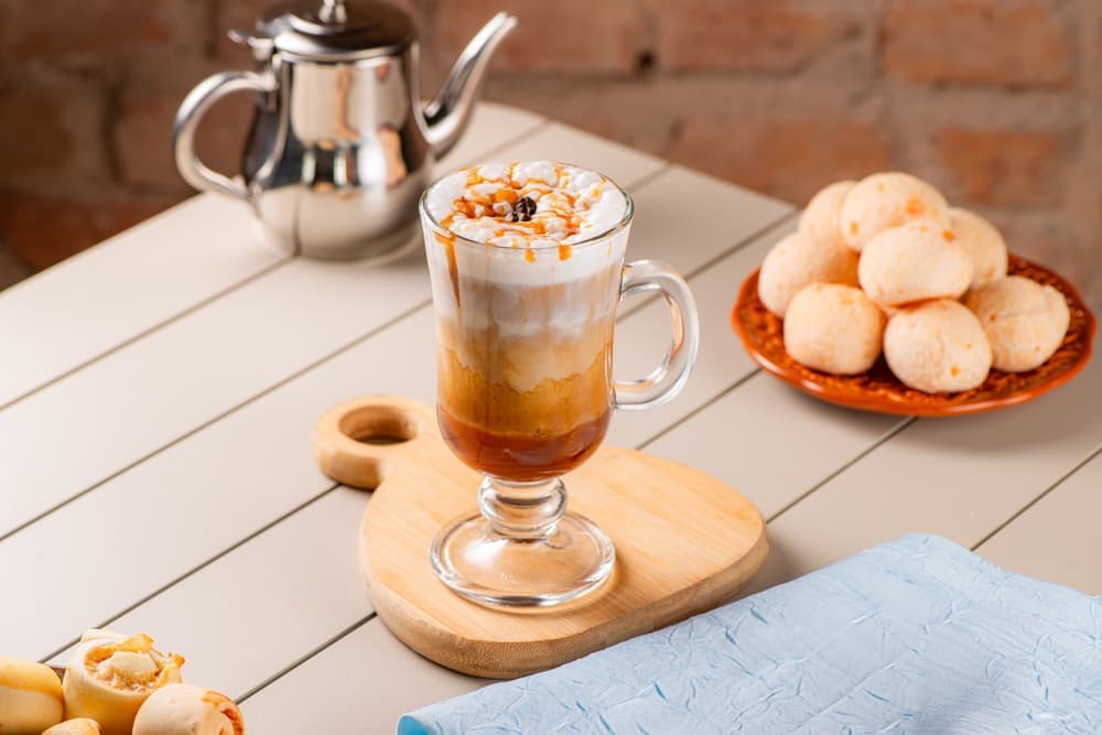 Café com leite em taça de vidro, com bule e pães de queijo.