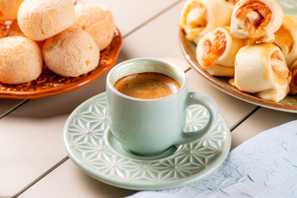 Café com leite em xícara verde, com enroladinhos e pães de queijo.