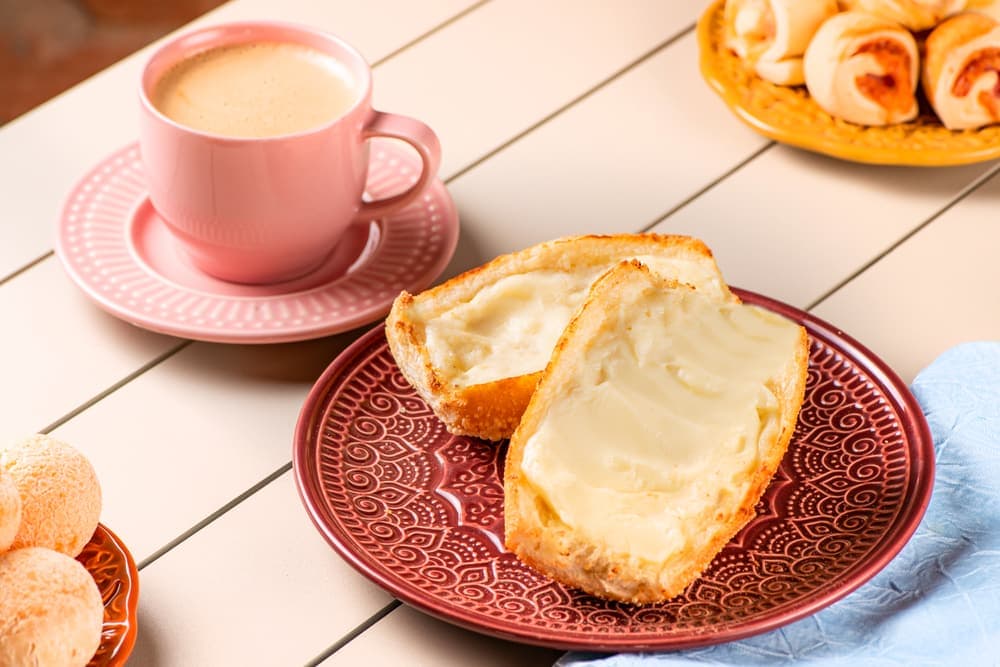 Pão francês na chapa com manteiga, com café em xícara rosa.