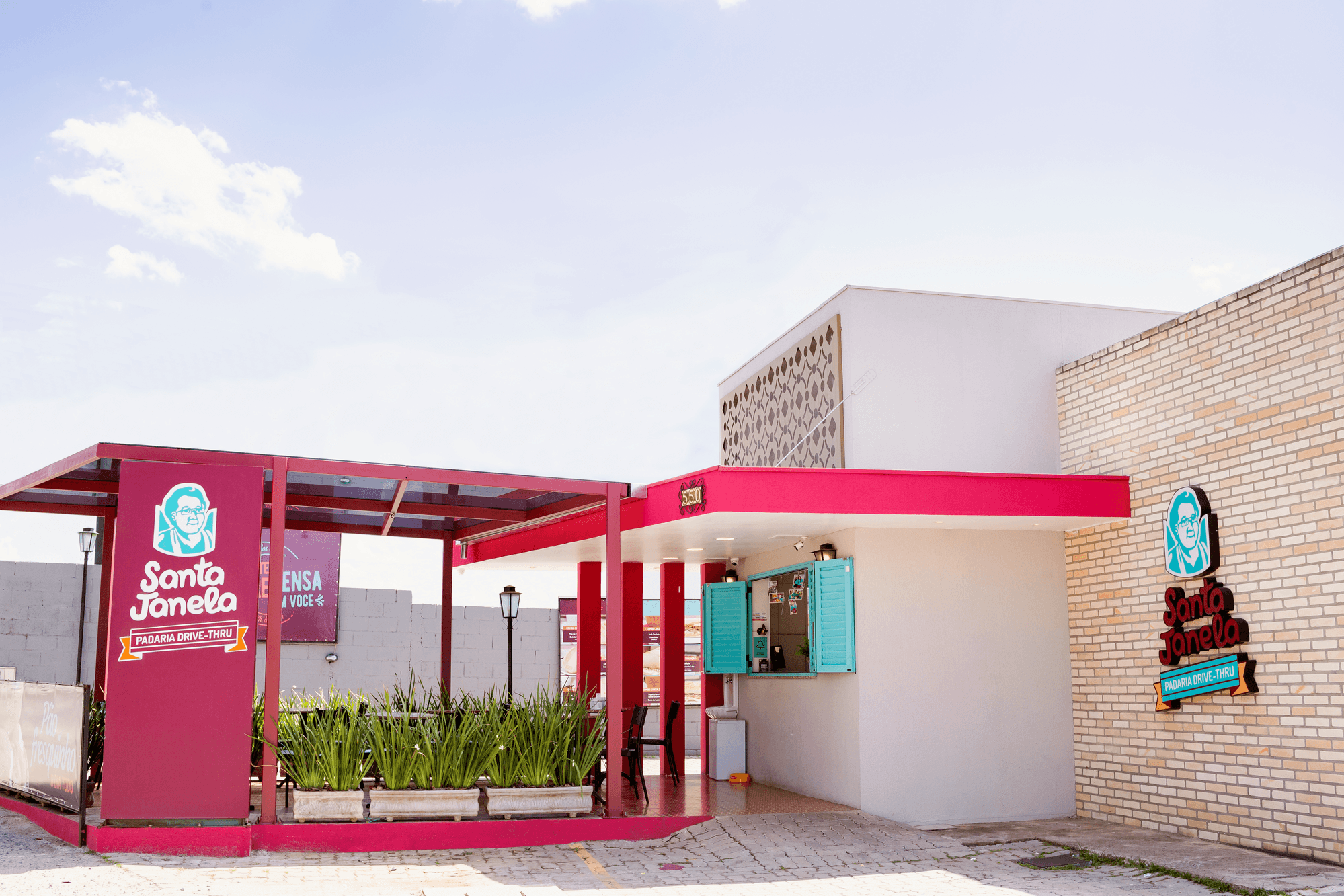 A unidade drive-thru da Santa Janela vista da rua: a marquise vermelha sobre a pista, a placa de padaria drive-thru e a janela de atendimento com as venezianas verdes abertas.